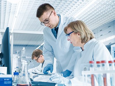 In Modern Laboratory Senior Female Scientist Has Discussion with Young Male Laboratory Assistant. He Shows Her Data Charts on a Clipboard, She Analyzes it and Enters It into Her Computer.