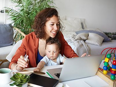 Attractive young dark skinned woman working at desk at home using laptop, holding baby on her lap. Portrait of smiling mother writing post on moms blog while her infant son playing with toy