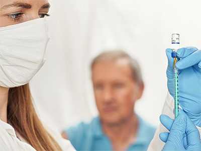 Young nurse with white cotton mouth virus face mask, injecting coronavirus vaccine (sticker is own design) into green syringe, blurred patient in background. Covid 19 cure concept